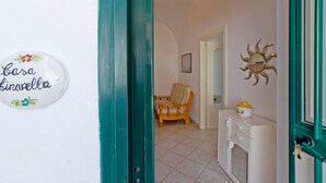 Um corredor com uma porta azul-esverdeada, uma placa com o dizer 'Casa Giravelle', uma cadeira de vime e um armário com um objeto amarelo em cima.