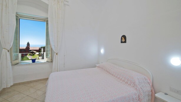 Quarto com cama, janela com cortinas, pequena decoração na parede e vista para edifícios e uma torre.