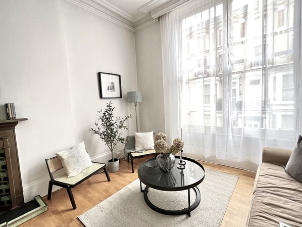 Un salon avec une table basse ronde, deux fauteuils, un canapé, une cheminée et une grande fenêtre ornée de rideaux légers.
