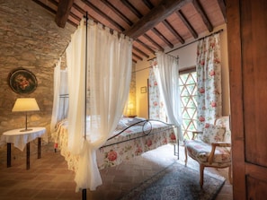 A bedroom with a four-poster bed, a wooden chair, a small table with a lamp, and a stone wall.