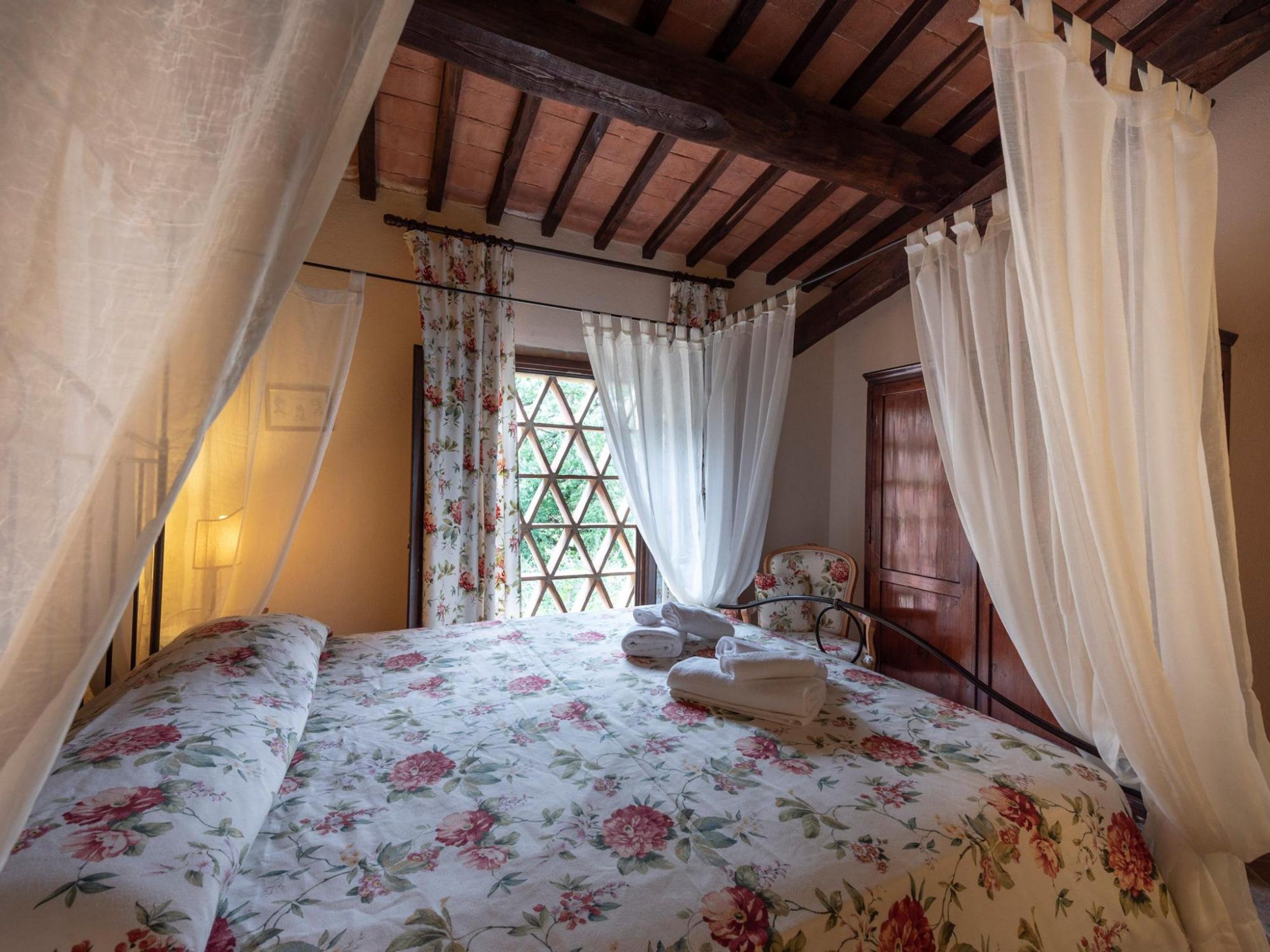 Un dormitorio con edredón estampado con motivos florales, cabecera de madera, ventana con diseño de celosía y armario de madera.