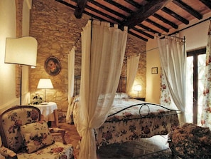 A bedroom with a stone wall, a canopy bed, a floral chair, and a bedside table with a lamp. - Apartment in Chianti With Pool ID 453 (Casole d'Elsa)