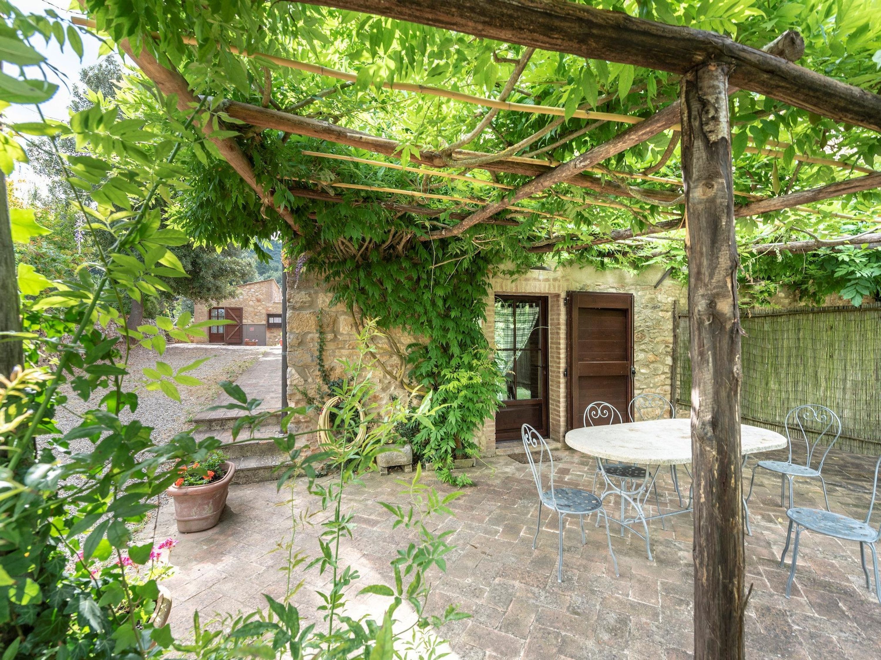 A rustic outdoor patio with a stone floor, a pergola covered with vines, and a table with metal chairs.