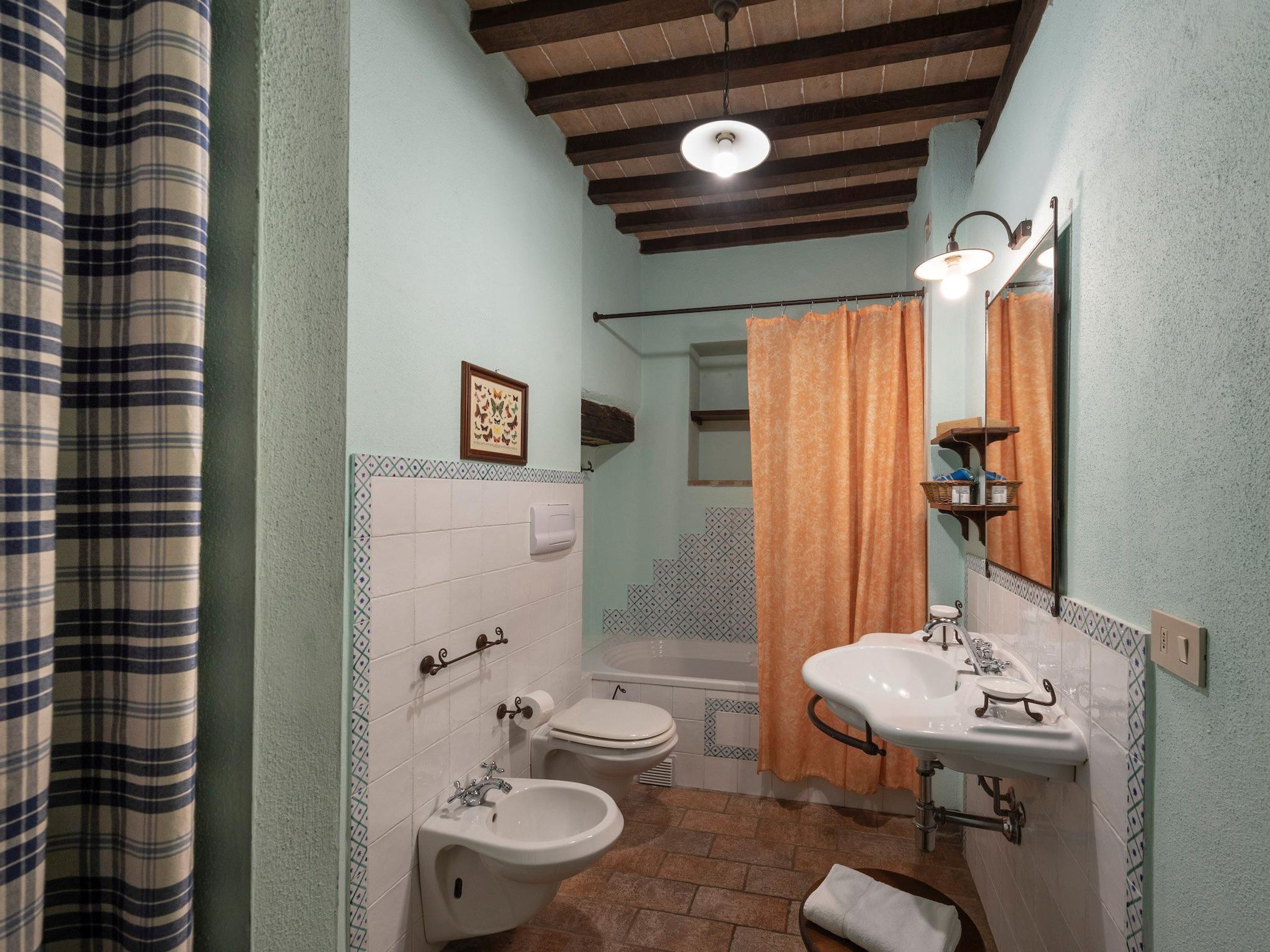 Un baño con inodoro, bidé y lavamanos. Hay una cortina de ducha, una toalla y un cuadro en la pared.