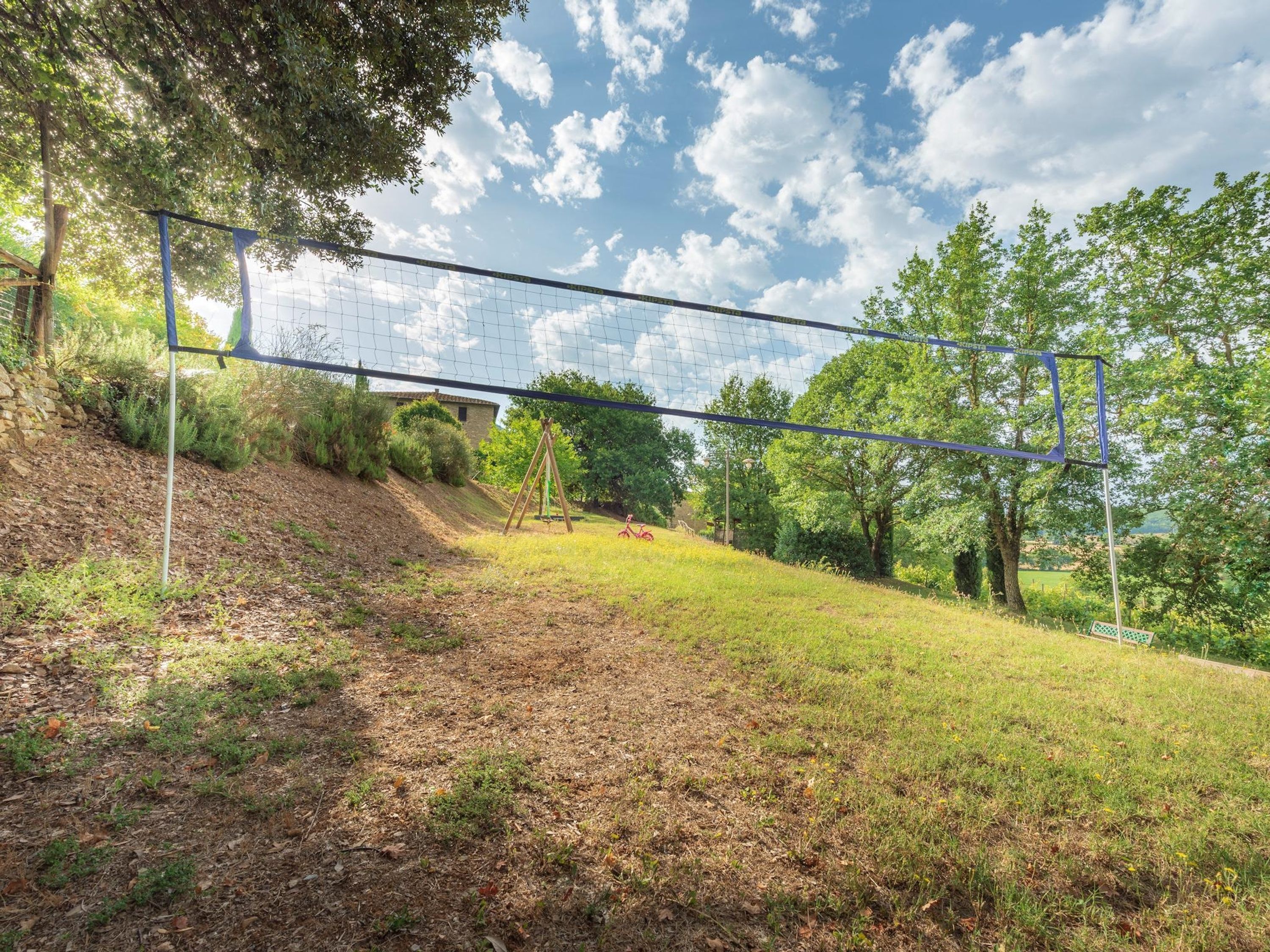 A tennis court with a net, surrounded by trees and a grassy slope.
