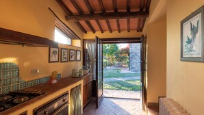 A kitchen with a stone floor, wooden beams, and a view of a garden through an open door. - Apartment in Chianti With Pool ID 455 (Casole d'Elsa)