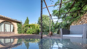 Piscina cubierta por cristal con una pérgola y zona de estar, rodeada de exuberante vegetación y un muro de piedra.