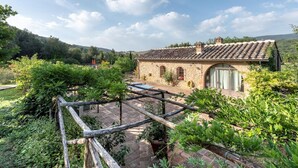 Un edificio de piedra con techo de tejas, rodeado de exuberante vegetación y una pérgola de madera.