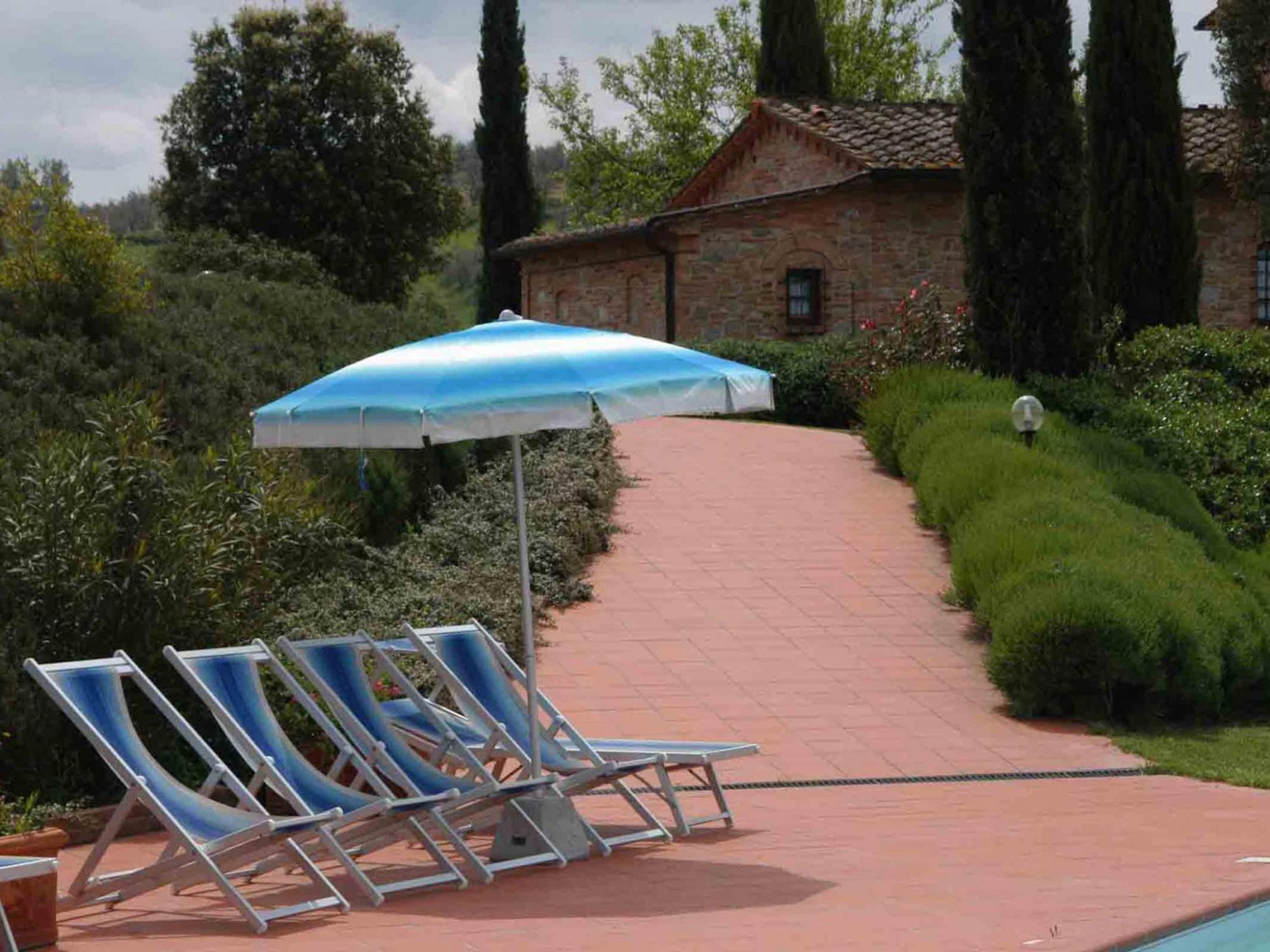 Ein Weg führt zu einem Poolbereich mit Liegestühlen und einem blauen Sonnenschirm.
