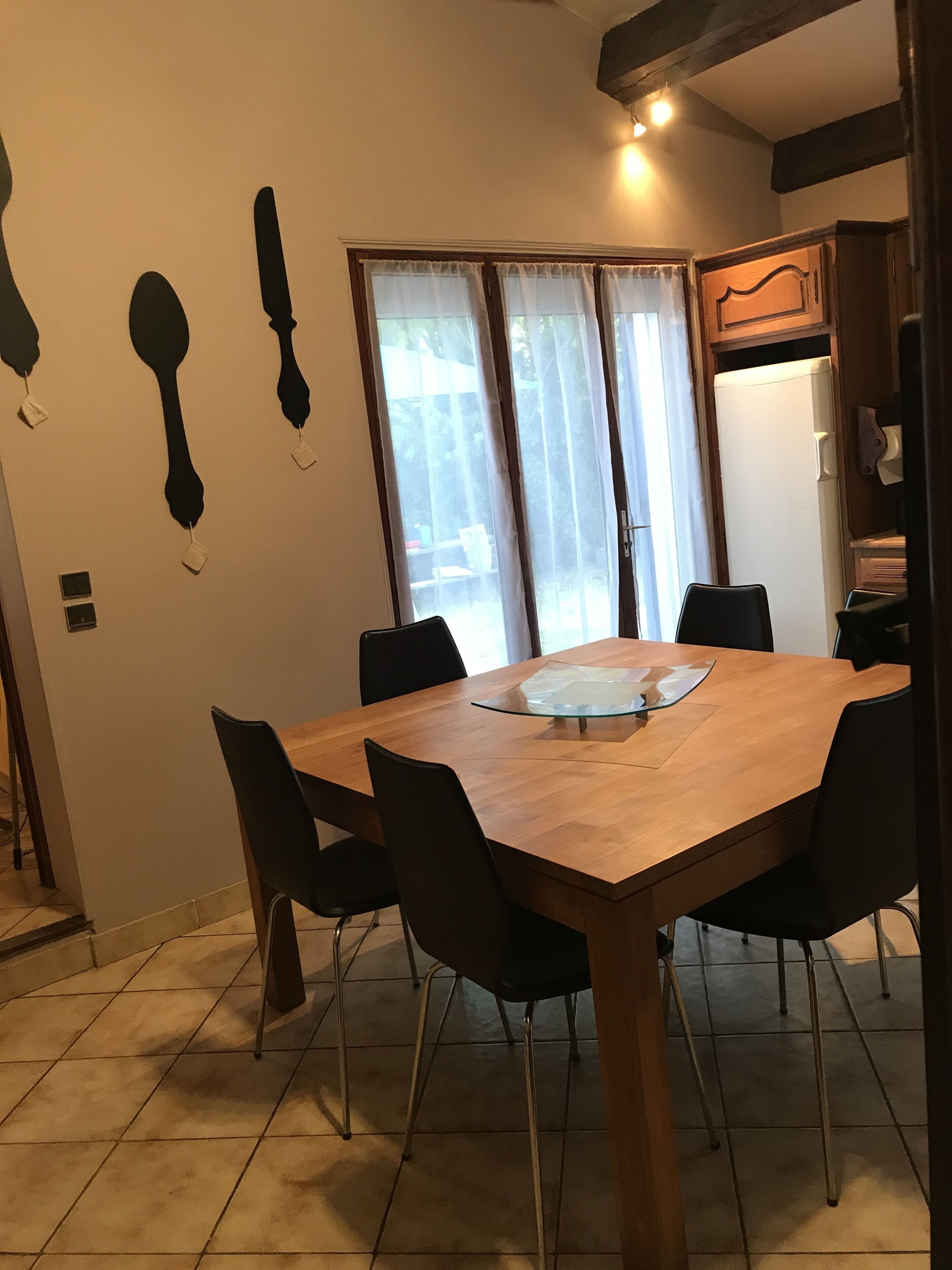 A dining area with a wooden table and black chairs, a window with curtains, and a refrigerator in the background.