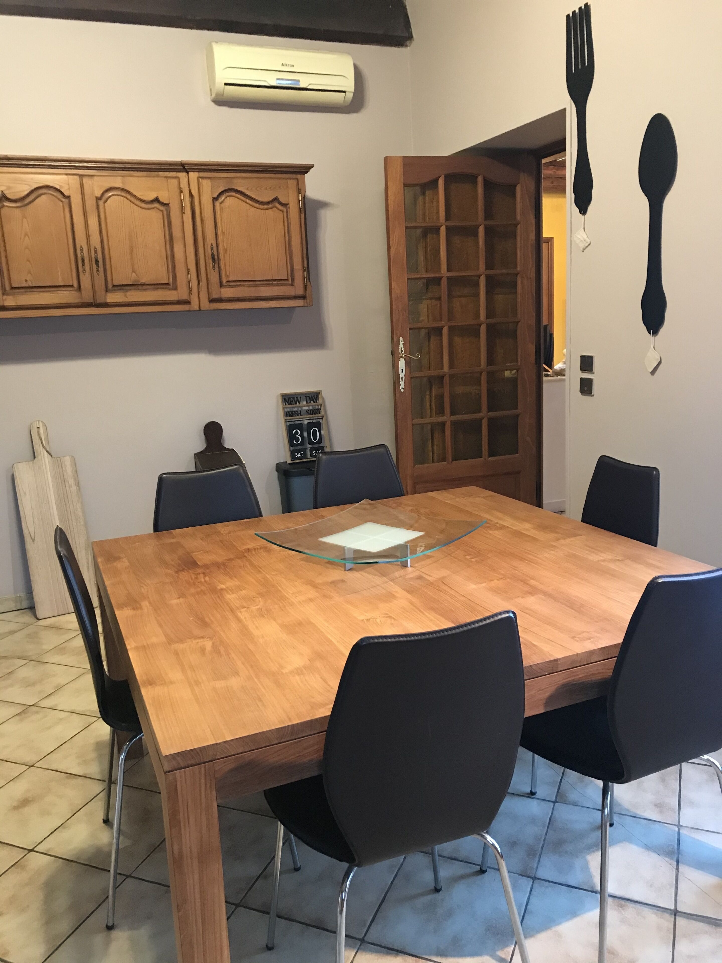 A dining area with a wooden table, black chairs, a glass-top center table, wooden cabinets, a door with glass panels, and wall-mounted utensils.