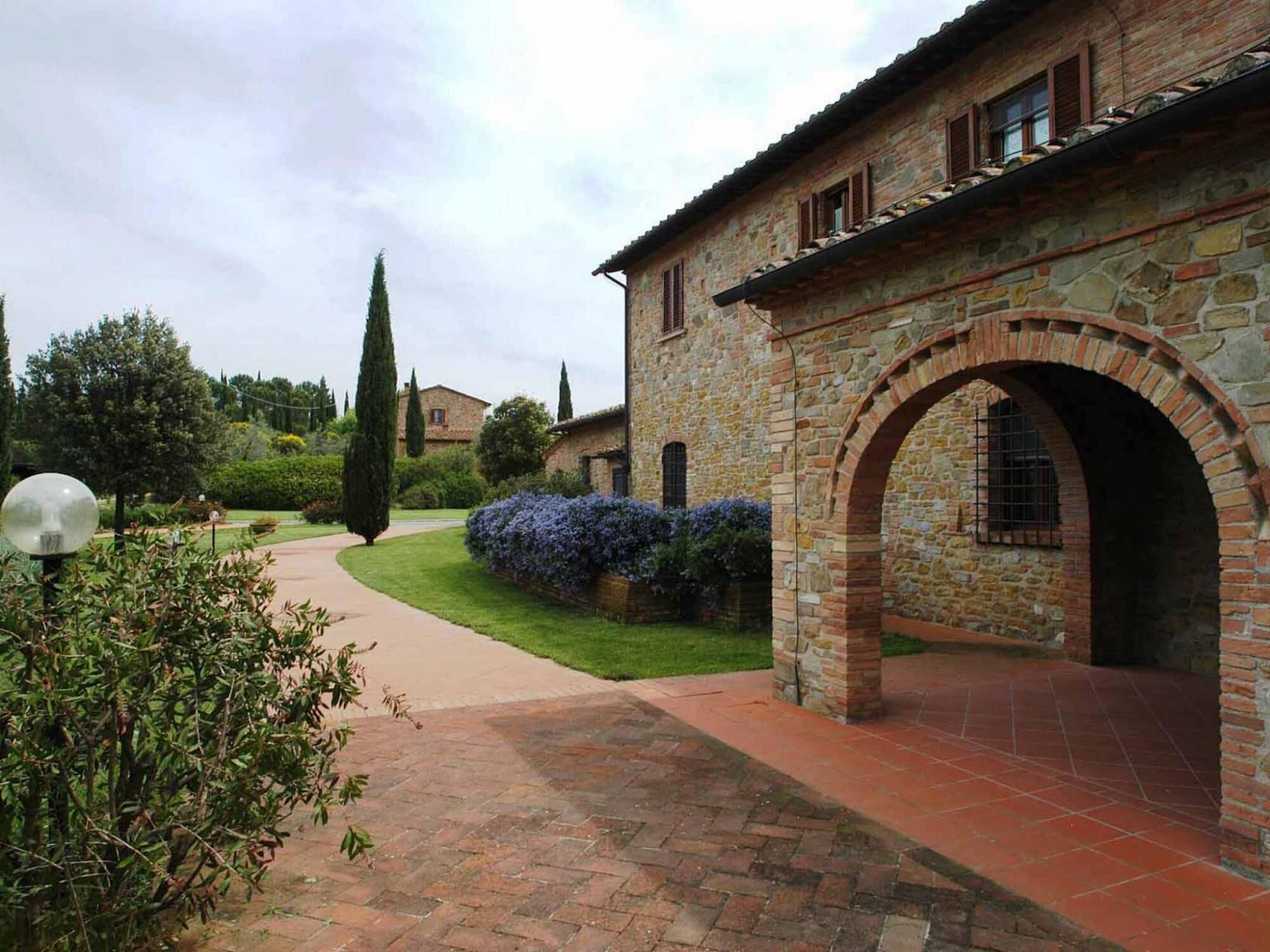 A stone building with an arched entrance, a paved pathway, and a well-maintained garden with various plants and a tall tree.