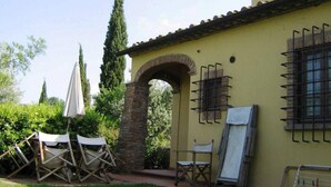 Un bâtiment à la façade jaune, doté d’une arche en pierre et d’une fenêtre avec des volets grillagés. On y trouve des chaises pliantes et un parasol blanc à l’extérieur.