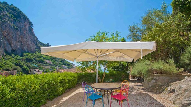 An outdoor patio with a round table and colorful chairs, a large umbrella, and a scenic view of a mountainous area with buildings in the background. - Relais San Basilio Convento - ID 3061 (Amalfi)