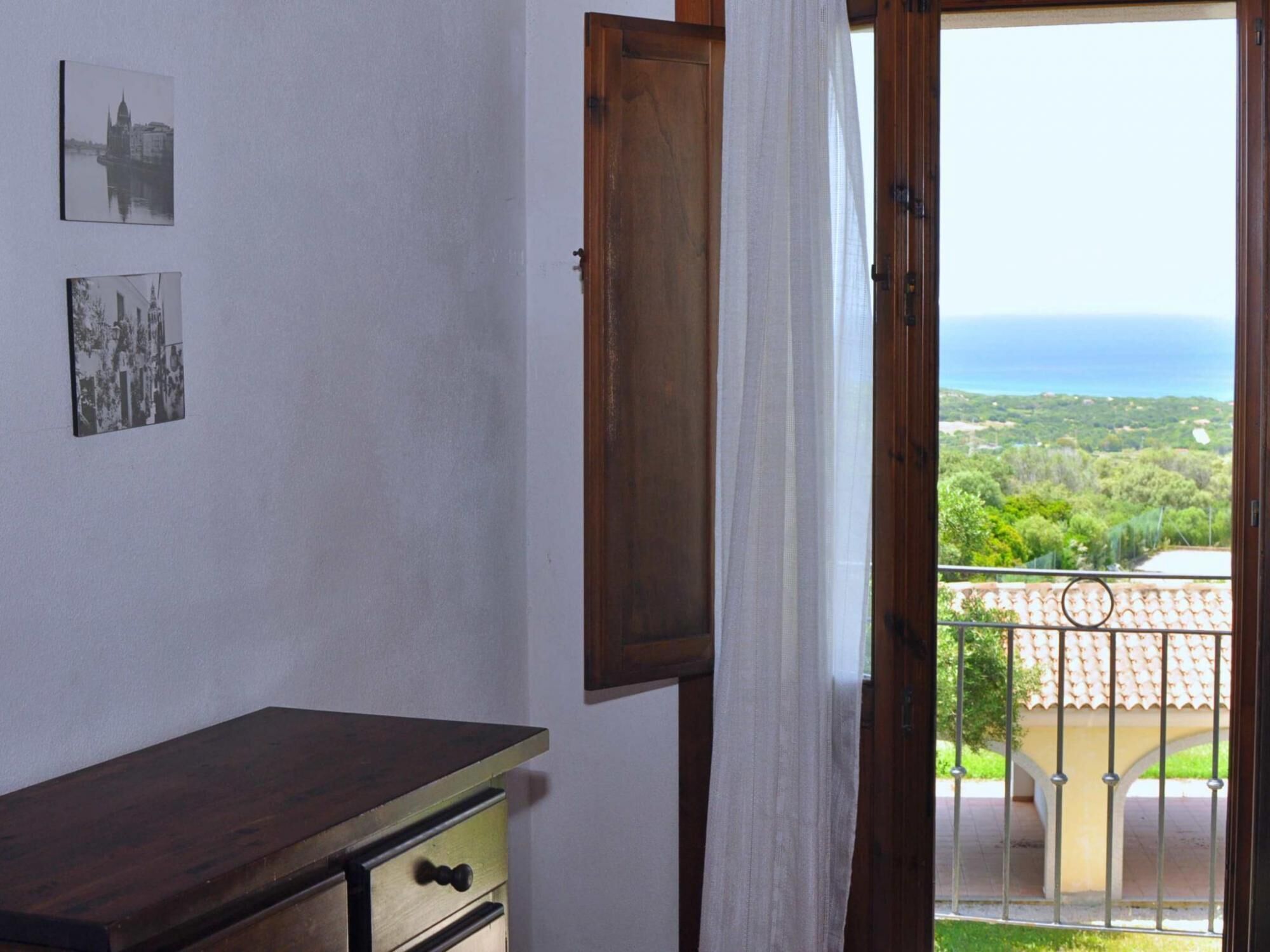 Ein Zimmer mit einem Holzkoffer, einer Tür zu einem Balkon mit Blick auf das Meer und viel Grün.