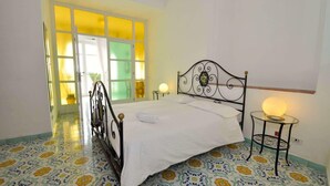 A bedroom with a bed, a bedside table with a lamp, a window with yellow-tinted glass, and a door leading to another room.
