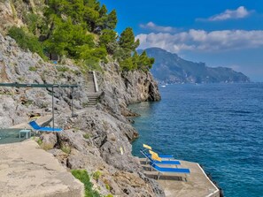 A rocky coastline with a staircase leading up, a small pool area with lounge chairs, and a view of the sea and distant mountains.