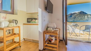A compact kitchen with a microwave, toaster, and a wooden shelf. A small table with books and a vase of flowers is near a sliding glass door leading to a balcony with a mountain view.