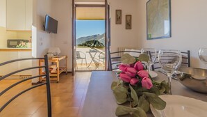 A dining area with a table set for two, a kitchen in the background, and a view of mountains through an open door.