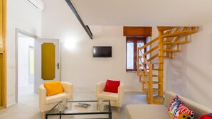 Un salon moderne avec une table basse en verre, deux fauteuils blancs ornés de coussins orange et rouges, un canapé beige et un escalier en colimaçon en bois.