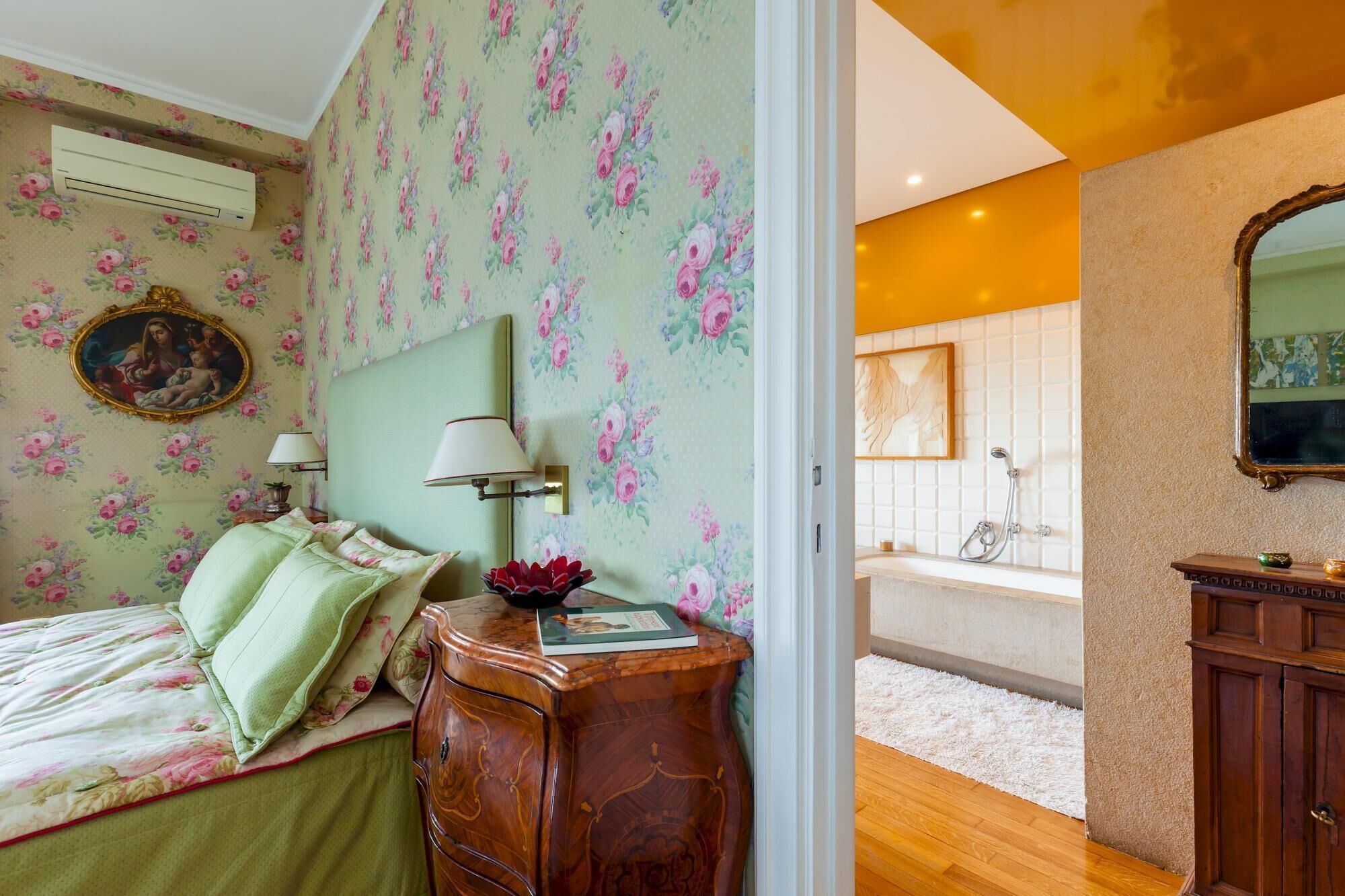 Un dormitorio con papel tapiz floral, una cama con ropa de cama verde, un tocador de madera y un baño con bañera y ducha.