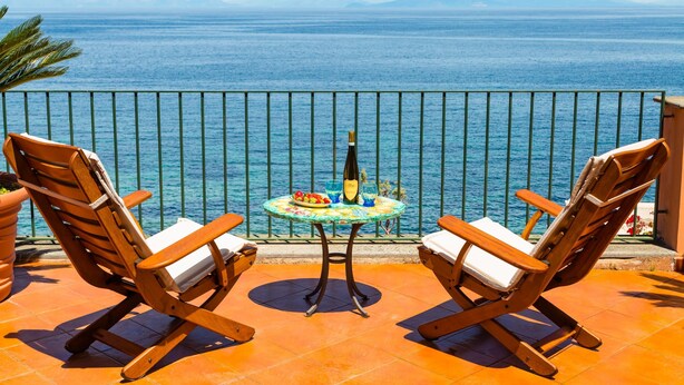 A terrace with two wooden chairs, a small table with a bottle and glasses, and a view of the sea. - Terrazza Morgera in Napoli (Casamicciola Terme)
