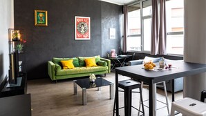 A modern living room with a green sofa, a coffee table, and a dining area with a table and chairs. There are two framed artworks on the wall and a window with curtains. - Le Loft Horloger (Besançon)