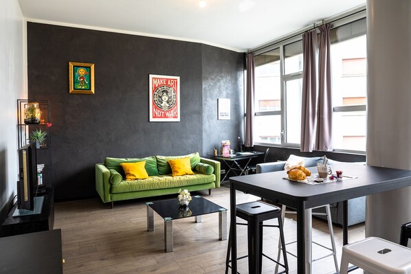 En moderne stue med en grøn sofa, et sofabord og et spiseområde med bord og stole. To indrammede kunstværker hænger på væggen, og et vindue er dækket af gardiner.