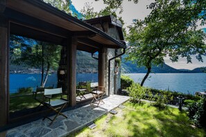 Une cabane moderne dotée de grandes fenêtres donnant sur un lac, d’un sol en pierre et d’un espace salon extérieur.