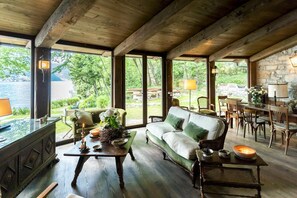 Un intérieur chaleureux et rustique, avec un plafond en bois, de grandes fenêtres donnant sur l’extérieur, un canapé vert et un coin repas avec des meubles en bois.
