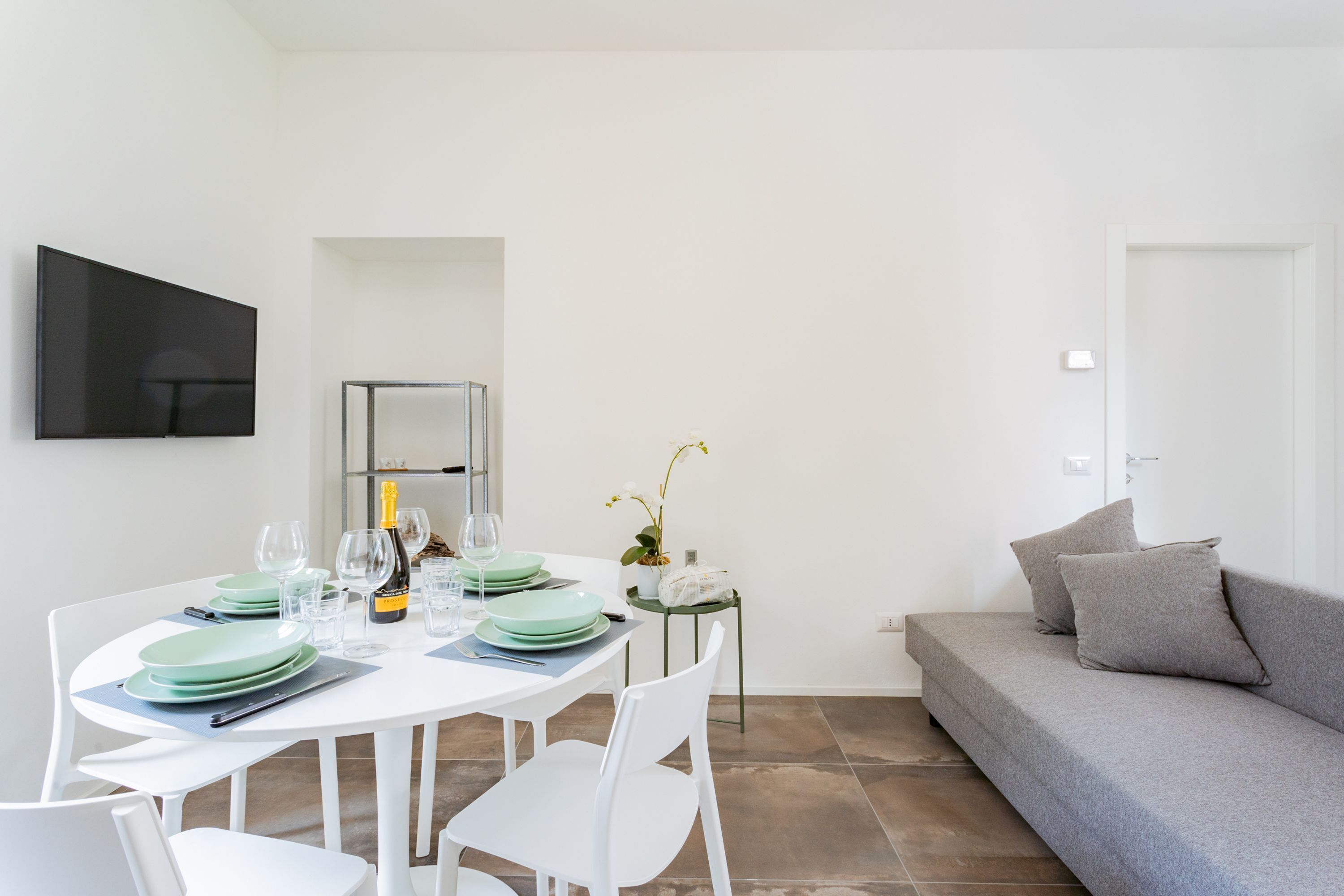Uma sala de estar moderna com uma mesa de jantar posta para quatro pessoas, um sofá cinzento e uma televisão de tela plana na parede.