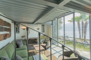 Ein großzügiger Wohnbereich mit einem grünen Sofa, einem Couchtisch und einem Kamin. Eine Treppe führt zu einem Balkon mit Blick auf Palmen und das Meer.