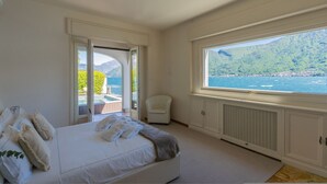 A bedroom with a large bed, a chair, and a view of the sea and mountains through a window. - Villa Bianca - Front Lake Pool (Sala Comacina)