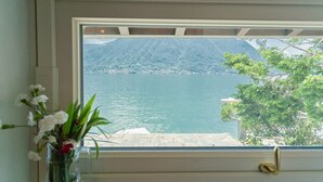 A window with a view of a calm body of water, a mountainous landscape, and a tree with green leaves. - Villa Bianca - Front Lake Pool (Sala Comacina)