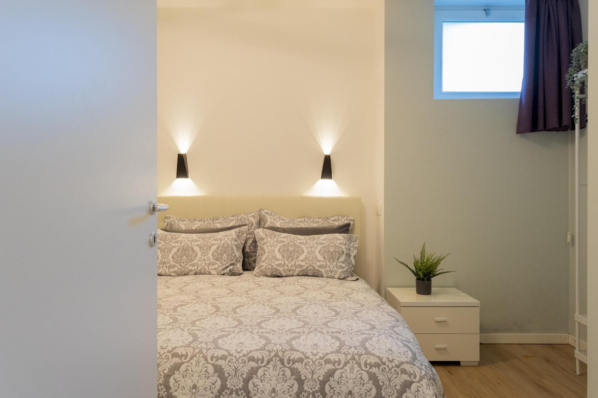 Quarto com uma cama, duas luminárias de cabeceira, mesa de cabeceira, janela e um vaso com planta.