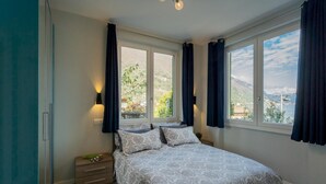 Ein Schlafzimmer mit einem Bett, zwei Fenstern, einem Nachttisch und Blick auf einen See und Berge.