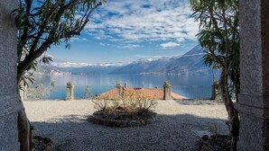 Blick auf einen See mit Bergen im Hintergrund, eine gepflasterte Fläche mit Fliesenbordur und ein Zaun, der einen Garten begrenzt.