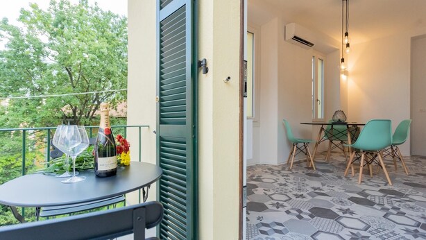 Ein Balkon mit Tisch für zwei Personen, einer Champagnerflasche und einem Glas. Innen im Gebäude befindet sich ein Essbereich mit Tisch und Stühlen.