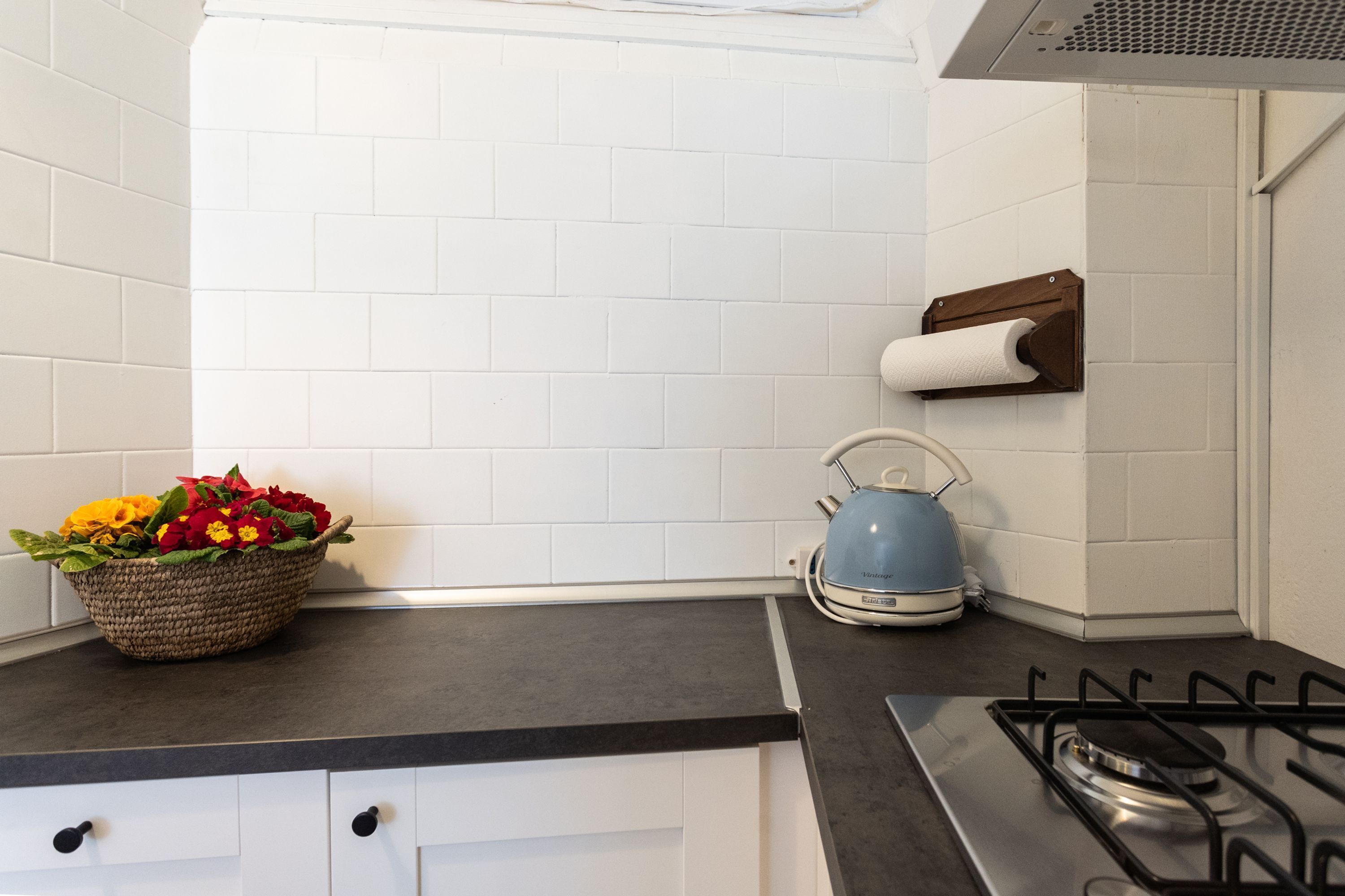 Una cucina con pareti in piastrelle bianche, un piano di lavoro nero, un fornello a gas, un bollitore e un cesto di fiori.