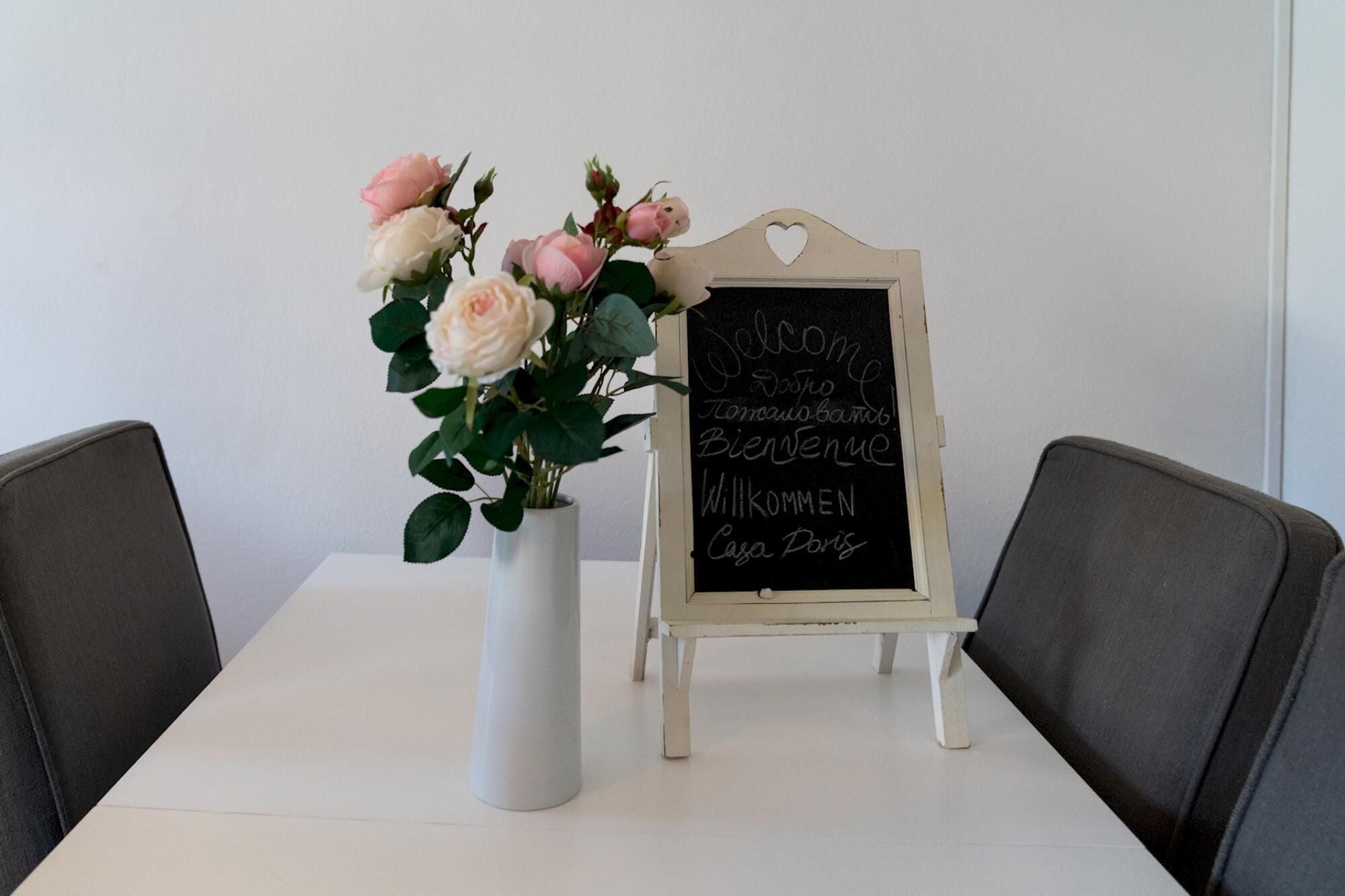 A white table with a white vase containing pink and white roses, and a small chalkboard with "Welcome" and "Bienvenue" written in multiple languages.