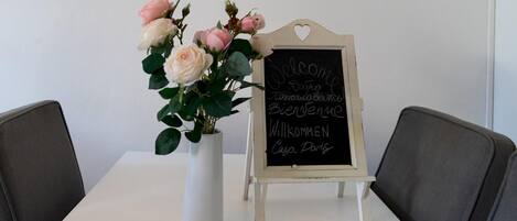 A white table with a white vase containing pink and white roses, and a small chalkboard with "Welcome" and "Bienvenue" written in multiple languages.
