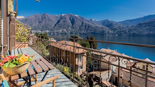 Ein Balkon mit Blick auf einen See und Berge, ein Tisch mit einer Blumenkorb und ein Geländer mit dekorativen Elementen.
