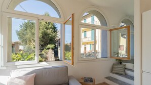A room with a sofa, a small table with a book, and a view of a garden and buildings through large windows. - Co-m341-sabb12a1 - Sunny Corner Near Bellagio (Tremezzina)