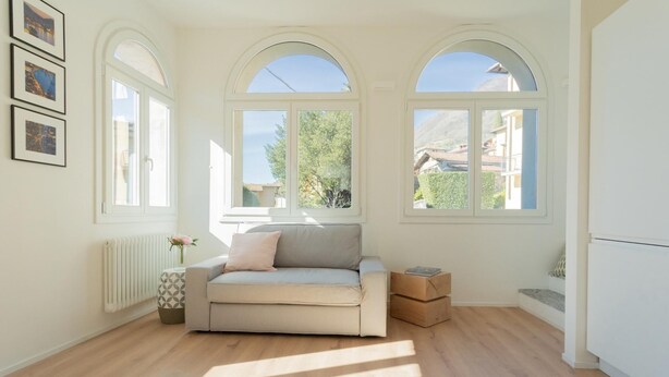 A bright room with a beige sofa, a radiator, and a window with a view of greenery and buildings. - Co-m341-sabb12a1 - Sunny Corner Near Bellagio (Tremezzina)