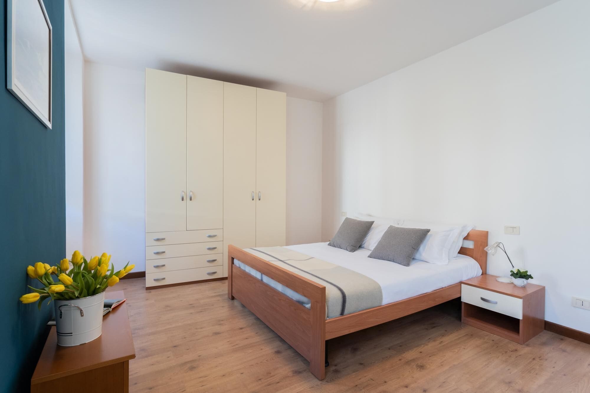 A bedroom with a bed, bedside table, nightstand, wardrobe, and a vase with flowers.
