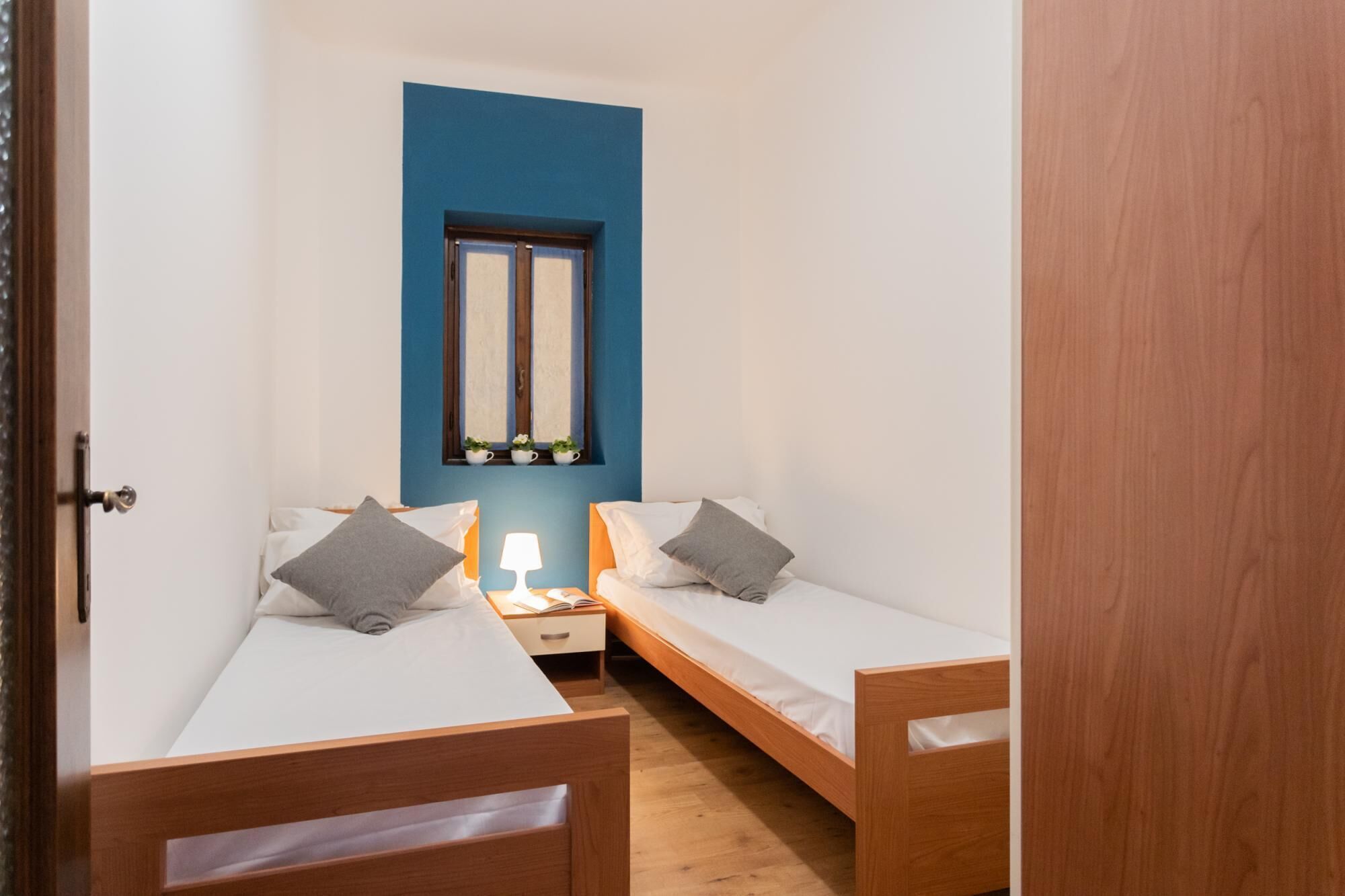 Una habitación pequeña y funcional con dos camas individuales, una mesita de noche con lámpara y una ventana con espejo arriba.