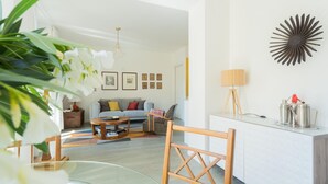Ein modernes Wohnzimmer mit Sofa, Couchtisch und Sesseln. Es gibt einen Essbereich mit einem Glastisch und Stühlen. Die Wand ist weiß, mit gerahmten Bildern und einem dekorativen Wandbild versehen.