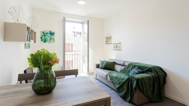 A modern living room with a wooden table, a sofa with a green blanket, and a large window with a view of buildings. - Co-c933-ggar24c4 - Central Green Apartment (Como)