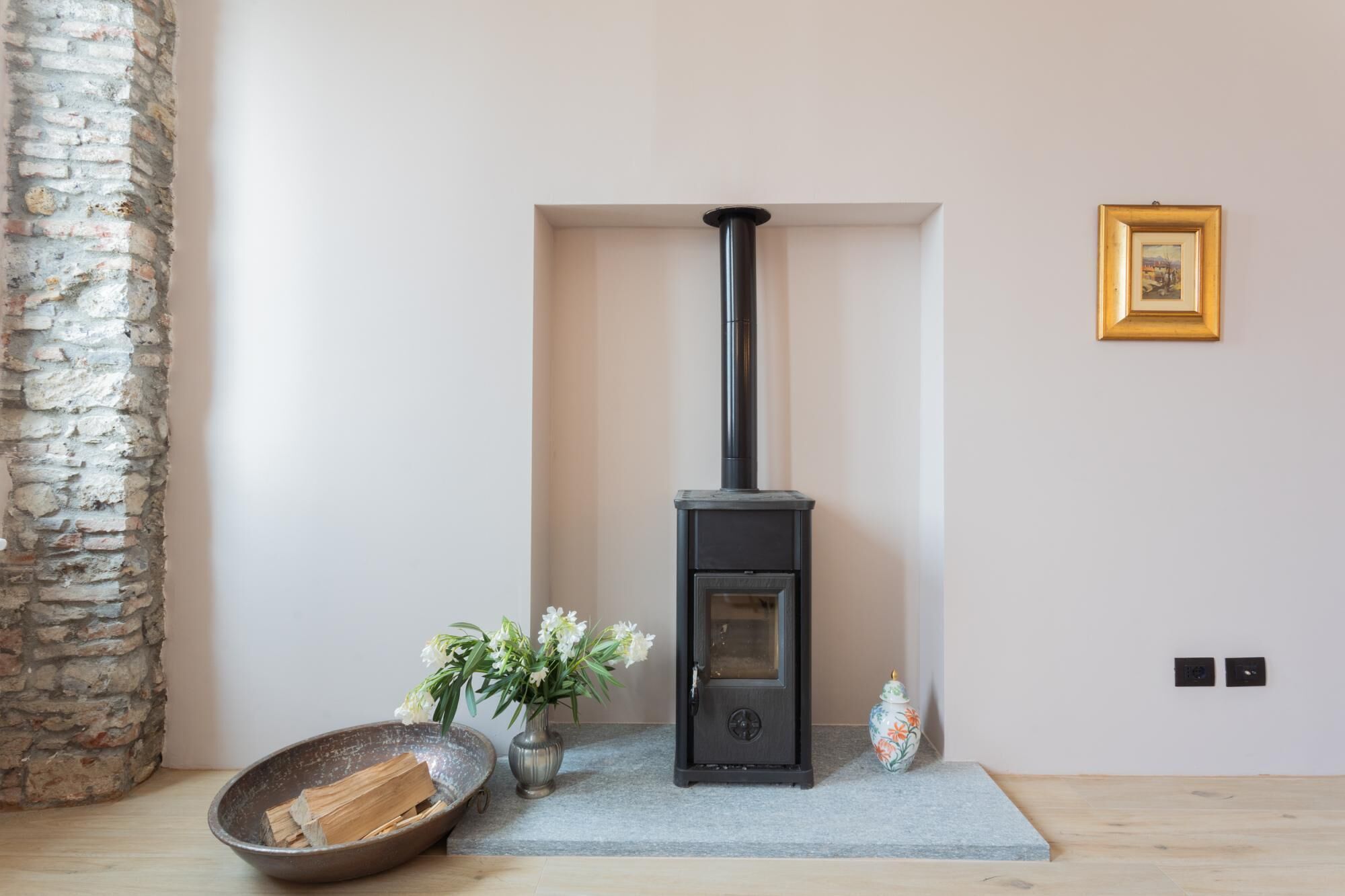 A room with a black wood-burning stove, a stone wall, a vase with flowers, and a framed picture on the wall.