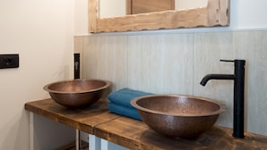 Une salle de bain avec deux vasques en cuivre posées sur un plan de travail en bois, un robinet noir et un meuble en bois surmonté d’un miroir.